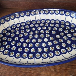 May include: An oval ceramic serving platter featuring a cream-colored base with a repeating pattern of blue, green, and brown concentric circles resembling peacock eyes. The rim is finished with a solid dark blue border and thin radiating lines.