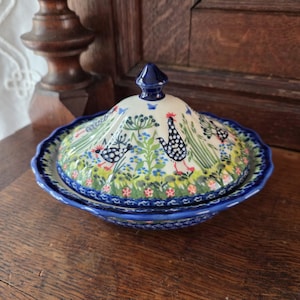 Op de afbeelding: Een ronde keramische botervloot met deksel, voorzien van een handgeschilderd patroon van zwart-wit gevlekte hanen, groen gras en kleine blauwe en rode bloemen. De schaal heeft een geschulpte rand en een diepblauwe afwerking aan de rand.