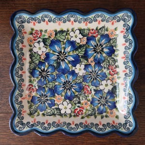 Op de afbeelding: Een vierkant keramieken bord met een geschulpte rand, voorzien van een crèmekleurige basis versierd met een levendig patroon van grote blauwe bloemen, kleinere witte bloesems, groene bladeren en kleine oranje accenten, afgewerkt met een donkerblauwe rand.