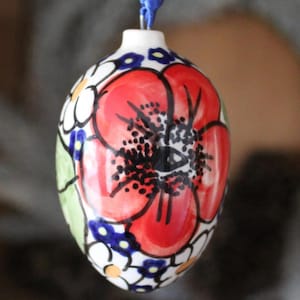 May include: A ceramic egg-shaped ornament featuring a hand-painted design of a large red poppy flower with black stamen details, surrounded by smaller white and yellow flowers, green leaves, and blue accents, suspended by a blue ribbon loop.