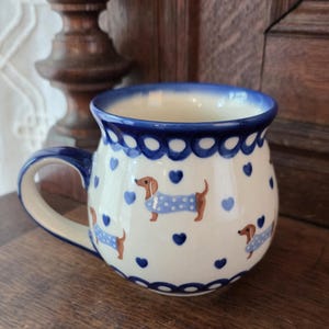 Puede incluir: Una taza de cerámica con cuerpo redondeado que presenta un patrón de perros salchicha marrones con suéteres de lunares azul claro, intercalados con pequeños corazones azul oscuro. El borde y el asa son azul oscuro, con un borde festoneado.