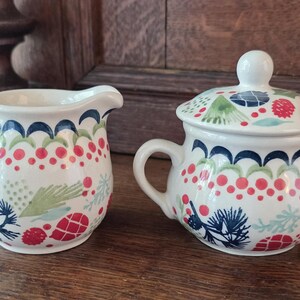 May include: A ceramic creamer and a lidded sugar bowl featuring a white base with a colourful pattern of red berries, green pine needles, dark blue pinecones, and scalloped blue lines. Both pieces have rounded bodies and curved handles.