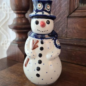 Może przedstawiać: Ceramiczna figurka bałwana z białym korpusem w niebieskie kropki, ciemnoniebieskim cylindrem z białymi okrągłymi akcentami, czerwonym nosem i czarnymi guzikami. Bałwan ma na sobie ciemnoniebieski szalik i trzyma w ręku brązową miotłę.