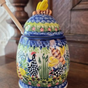 May include: A ceramic honey pot with a lid featuring a yellow bee handle. The pot is decorated with a colorful pattern of black and white spotted chickens, yellow bees, green grass, and blue flowers. A wooden honey dipper rests inside the pot.