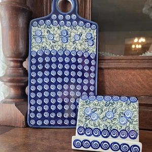 Könnte beinhalten: Ein rechteckiges Servierbrett aus Keramik mit Griffloch und ein passender Keramikständer, beide mit einem dunkelblau-weißen Spiralmuster sowie einem hellgrün-blauen Blumenmotiv auf cremefarbenem Hintergrund verziert.