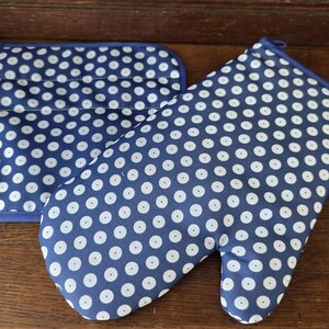 Könnte beinhalten: Ein blauer Ofenhandschuh und ein quadratischer Topflappen, beide mit einem sich wiederholenden weißen Punktmuster. Der Ofenhandschuh hat einen Daumenbereich und eine Aufhängeschlaufe, während der Topflappen quadratisch mit abgerundeten Ecken ist.