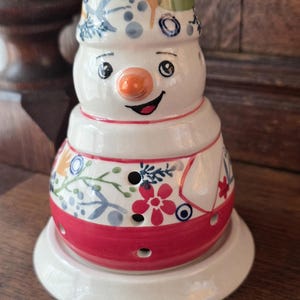 Peut inclure: Une figurine de bonhomme de neige en céramique avec une base blanche ornée d'un motif floral coloré en rouge, bleu, vert et orange. Le bonhomme de neige a un nez en carotte orange peint, des yeux noirs et une bouche souriante.