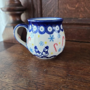 May include: A ceramic mug with a rounded body and a handle, featuring a cream-colored base decorated with blue snowmen, blue snowflakes, red and white candy canes, and small yellow stars, finished with a blue scalloped pattern at the rim.