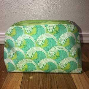 Pode incluir: Uma bolsa verde e azul com zíper, com um padrão de ondas e o texto "I see waves and a sheltered cove" repetido no tecido.