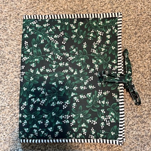 May include: A dark green fabric item with a white floral pattern and a black and white striped border. A matching fabric tie is attached to the side. The item is likely a journal or notebook cover.