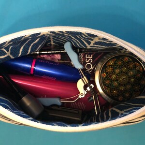 Puede incluir: Una bolsa de cremallera a rayas blancas y negras con un forro estampado en azul y blanco. La bolsa est&aacute; abierta y llena de varios art&iacute;culos de maquillaje y belleza, incluyendo m&aacute;scara de pesta&ntilde;as, delineador de ojos, un rizador de pesta&ntilde;as y un peque&ntilde;o espejo redondo.
