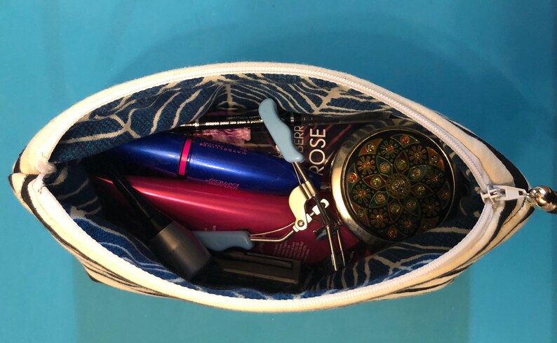 Puede incluir: Una bolsa de cremallera a rayas blancas y negras con un forro estampado en azul y blanco. La bolsa est&aacute; abierta y llena de varios art&iacute;culos de maquillaje y belleza, incluyendo m&aacute;scara de pesta&ntilde;as, delineador de ojos, un rizador de pesta&ntilde;as y un peque&ntilde;o espejo redondo.