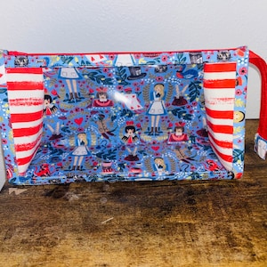 Könnte beinhalten: Eine blaue und rote Reißverschlusstasche mit einem transparenten Plastikfenster. Die Tasche ist mit einem fantasievollen Muster von Alice im Wunderland-Charakteren und einem roten und weißen gestreiften Stofffutter verziert.