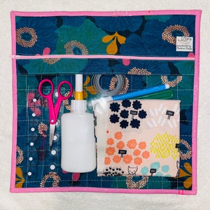 Puede incluir: Una bolsa de tela rosa con cierre de cremallera, llena de suministros de artesanía. La bolsa tiene un patrón floral en tonos de azul, verde y dorado. Los suministros incluyen un par de tijeras rosas, una botella de pegamento blanco, un rollo de cinta transparente, un marcador azul y un trozo de tela con un patrón colorido.