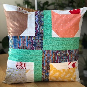 Op de afbeelding: Een vierkant kussen met een patchwork-ontwerp met verschillende stoffen in tinten bruin, groen, oranje, geel en rood. Het kussen is gemaakt van katoen en heeft een witte achtergrond.