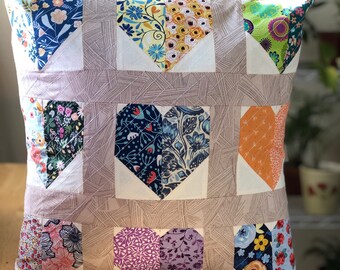 Peut inclure: Coussin carré décoratif avec un motif patchwork en forme de cœurs. Les cœurs sont faits de divers tissus à motifs floraux dans des tons de bleu, jaune, orange et violet. Le fond est marron clair avec un motif linéaire subtil.