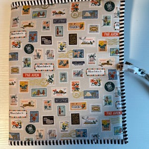 Könnte beinhalten: Ein mit Stoff bezogenes Notizbuch mit hellgrauem Hintergrund, verziert mit farbenfrohen Briefmarkenmotiven. Die Kanten sind mit einem schwarz-weiß gestreiften Rand versehen. Eine weiße Kordel ist an der Seite befestigt.