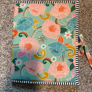 Könnte beinhalten: Ein floral bedrucktes Notizbuch mit türkisblauem Hintergrund und großen rosa und blauen Blumen. Der Einband hat einen schwarz-weiß gestreiften Rand und einen passenden Krawattenverschluss. Das Design umfasst Regenbogen, Schmetterlinge und andere florale Elemente.