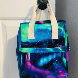 May include: A blue, green, and purple backpack with a white aurora borealis print. The backpack has two white straps and a zipper with a small tassel.