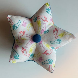 Puede incluir: Una almohada blanca en forma de estrella con un botón azul en el centro. La almohada está cubierta de una tela con un patrón repetitivo de contornos de manos de colores.