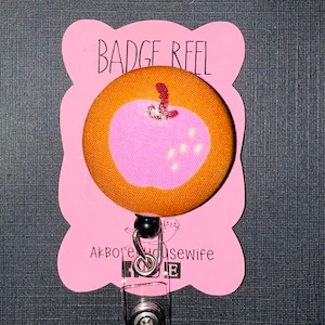 Pode incluir: Um carretel de crachá rosa e marrom com um design de maçã. O carretel tem um cordão retrátil de plástico transparente e um clipe de metal preto. O texto "Badge Reel" está impresso no cartão rosa.