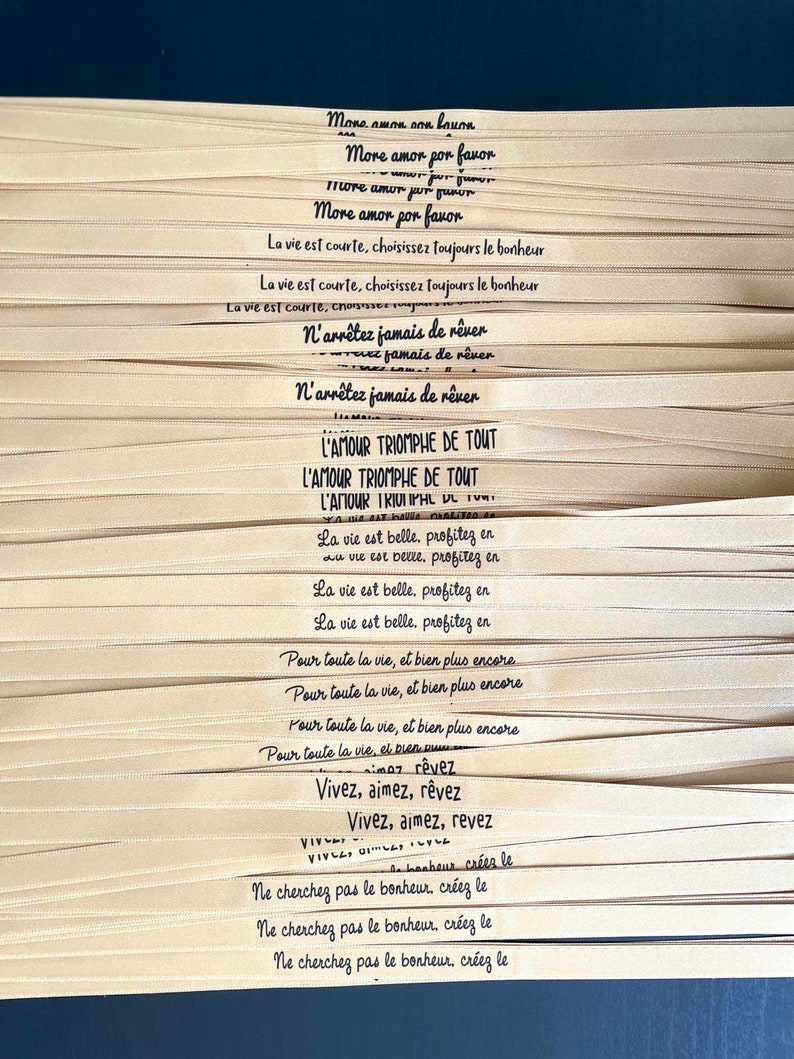 May include: Close-up of several beige ribbons with black script. The ribbons feature inspirational phrases in multiple languages, including "More amor por favor" and "L'amour triomphe de tout."