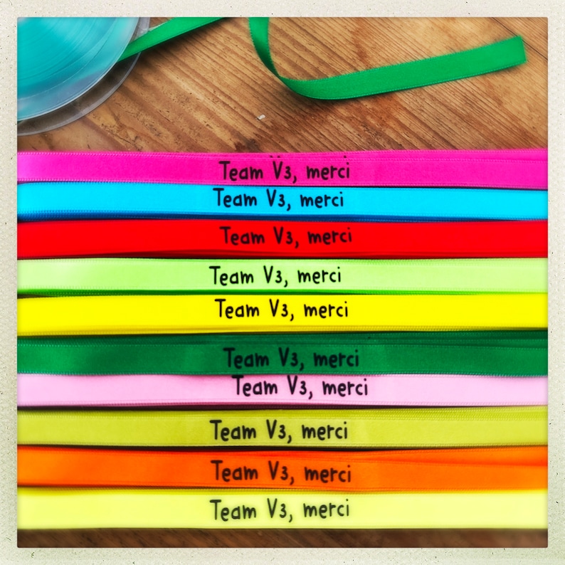 May include: A collection of colorful ribbons, each displaying the text "Team V3, merci" in black. The ribbons are in various shades, including pink, blue, red, green, yellow, and orange. A roll of ribbon and a green ribbon are visible in the background.
