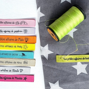 Peut inclure: Un ensemble de huit étiquettes en tissu colorées avec le texte "Les p'tites affaires de" suivi d'un nom et d'une petite icône. Les étiquettes sont disposées verticalement sur un fond blanc.