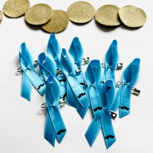 Peut inclure: Plusieurs épingles à ruban bleu clair, chacune ornée d'une petite moustache noire. Les épingles sont fixées avec des épingles de sûreté argentées et disposées sur une surface blanche, avec des confettis circulaires dorés au-dessus.