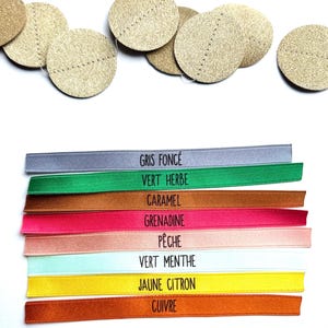 May include: A collection of gold-colored, circular embellishments with stitched details, alongside colorful ribbons. The ribbons are labeled with French color names: Gris Fonc&eacute;, Vert Herbe, Caramel, Grenadine, P&ecirc;che, Vert Menthe, Jaune Citron, and Cuivre.