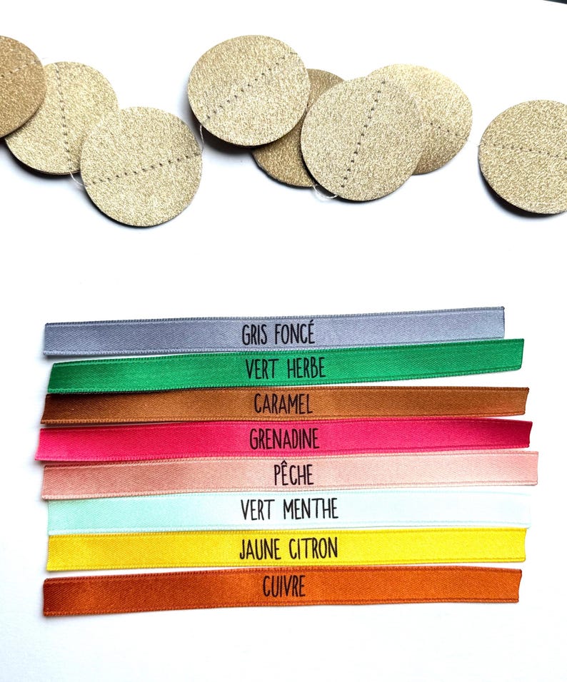 May include: A collection of gold-colored, circular embellishments with stitched details, alongside colorful ribbons. The ribbons are labeled with French color names: Gris Fonc&eacute;, Vert Herbe, Caramel, Grenadine, P&ecirc;che, Vert Menthe, Jaune Citron, and Cuivre.