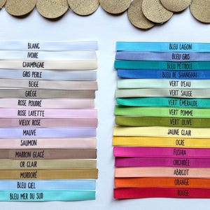 May include: A selection of colorful ribbons, each labeled with a different color name in French. The ribbons are stacked in two columns, with a variety of pastel and vibrant hues. Gold-colored circles are arranged at the top.