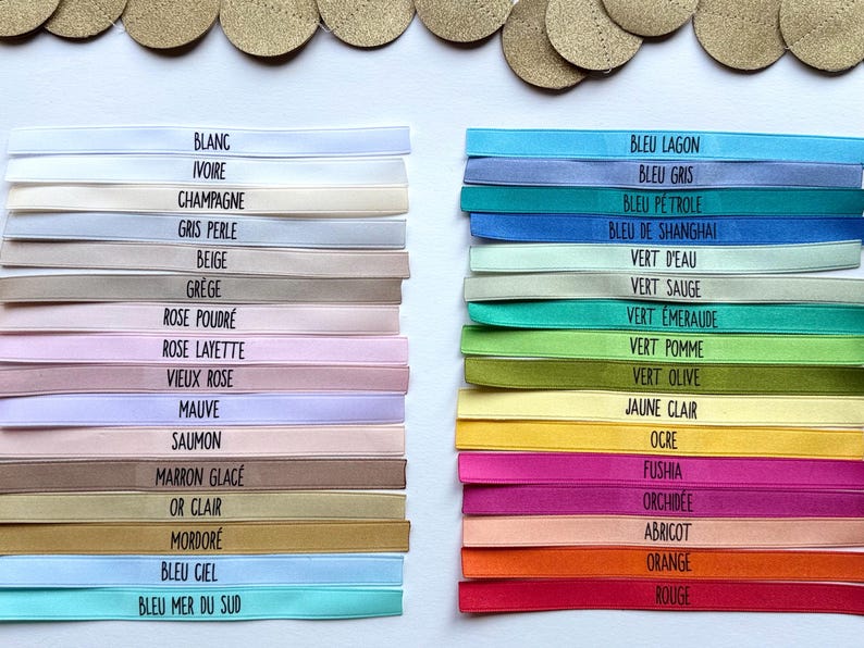 May include: A selection of colorful ribbons, each labeled with a different color name in French. The ribbons are stacked in two columns, with a variety of pastel and vibrant hues. Gold-colored circles are arranged at the top.