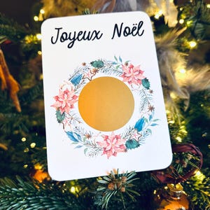 May include: A white card with the text 'Joyeux Noël' in black cursive font. The card features a watercolor wreath with pink flowers, green leaves, and gold stars. The center of the wreath is a gold circle.