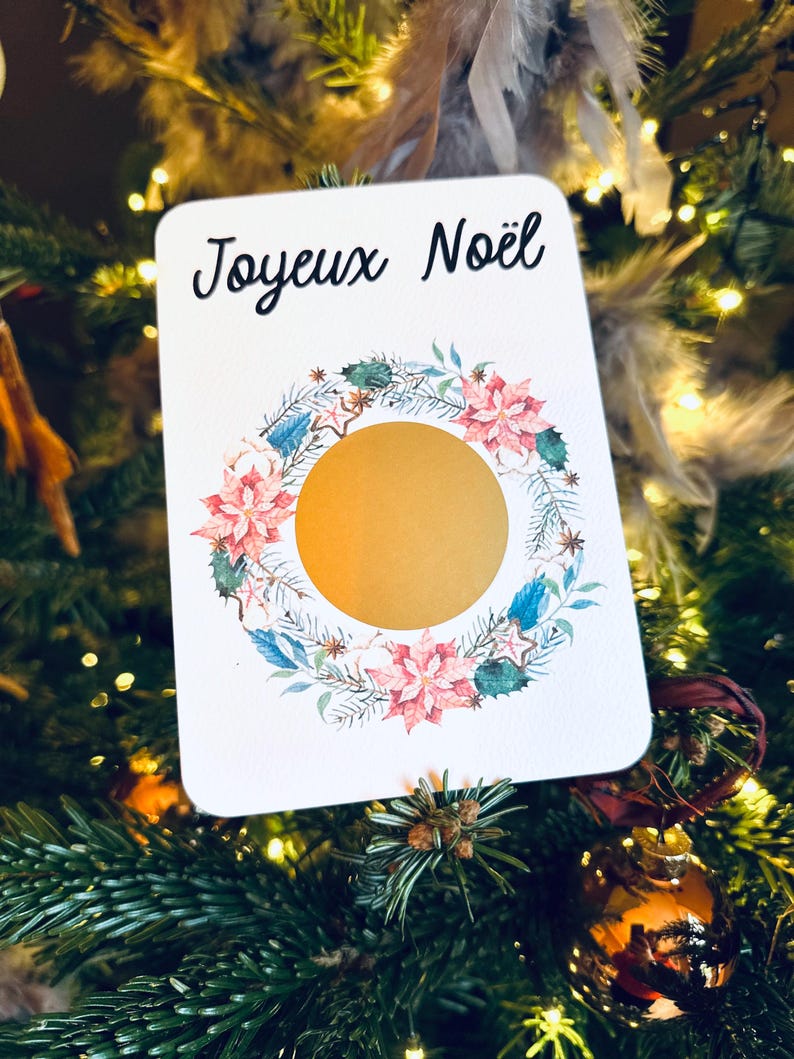 May include: A white card with the text 'Joyeux Noël' in black cursive font. The card features a watercolor wreath with pink flowers, green leaves, and gold stars. The center of the wreath is a gold circle.