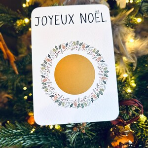 May include: A white card with the text 'JOYEUX NOËL' in black lettering. The card has a gold circle in the center surrounded by a wreath of green leaves, red berries, and pine cones.