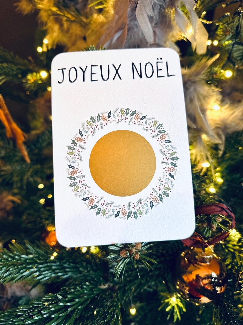 May include: A white card with the text 'JOYEUX NOËL' in black lettering. The card has a gold circle in the center surrounded by a wreath of green leaves, red berries, and pine cones.