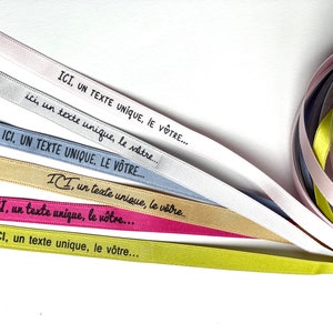 May include: Assorted satin ribbons in shades of pink, silver, blue, gold, and green. Each ribbon displays the text "ICI, un texte unique, le v&ocirc;tre..." in black. A roll of ribbons is visible in the background.