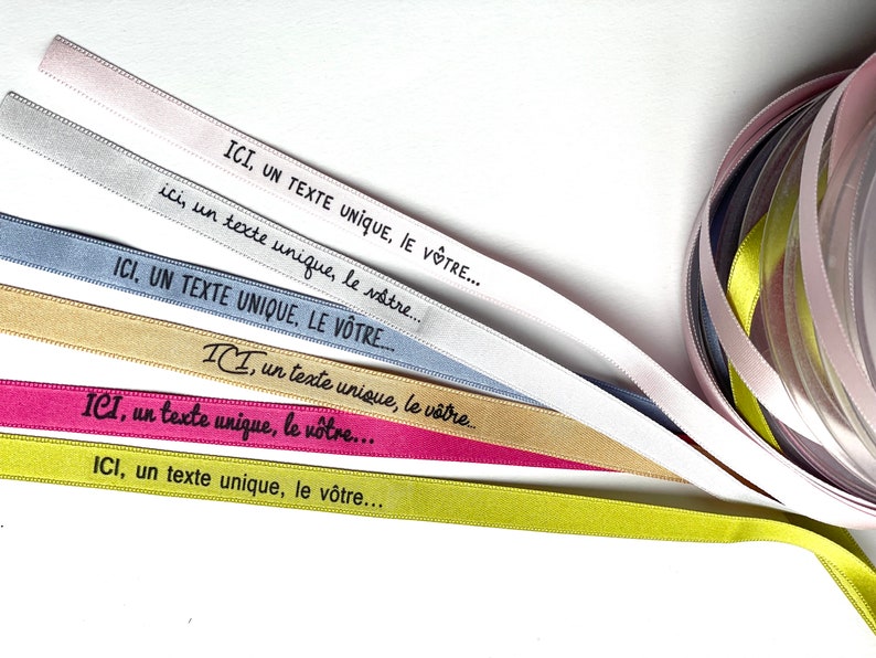 May include: Assorted satin ribbons in shades of pink, silver, blue, gold, and green. Each ribbon displays the text "ICI, un texte unique, le v&ocirc;tre..." in black. A roll of ribbons is visible in the background.