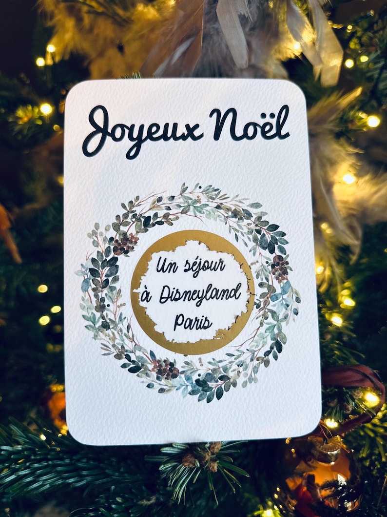 May include: A white card with a gold circle in the center. The card has a green wreath around the circle. The text on the card reads 'Joyeux Noël' and 'Un séjour à Disneyland Paris'.