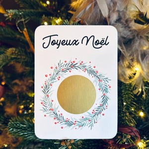 May include: A white card with the text 'Joyeux Noël' in black cursive writing. There is a gold circle in the center of the card surrounded by a green wreath with red berries.