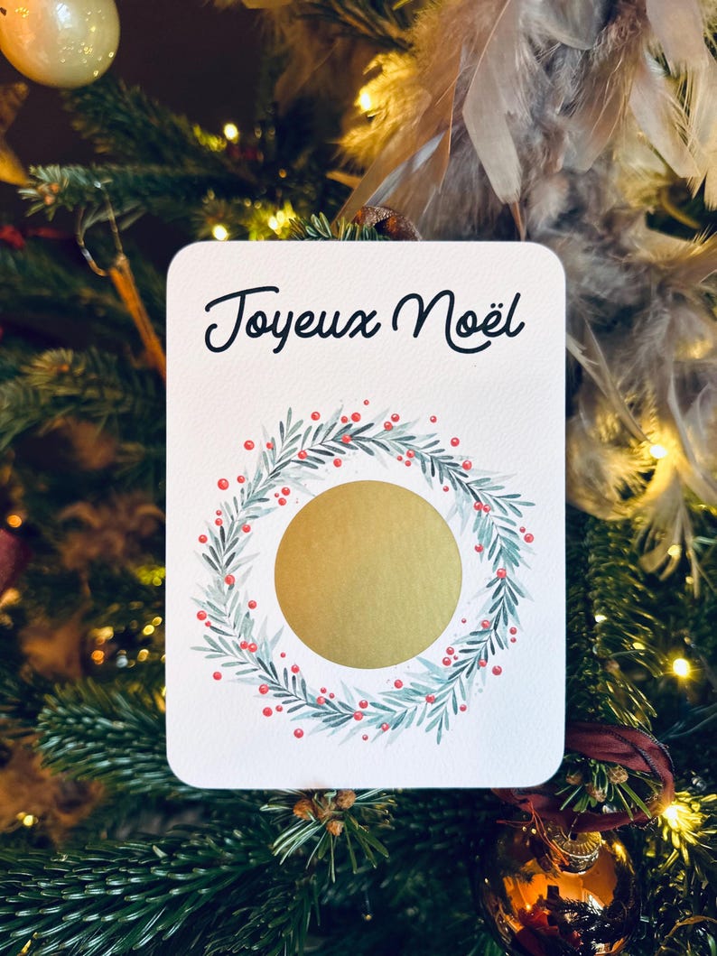 May include: A white card with the text 'Joyeux Noël' in black cursive writing. There is a gold circle in the center of the card surrounded by a green wreath with red berries.