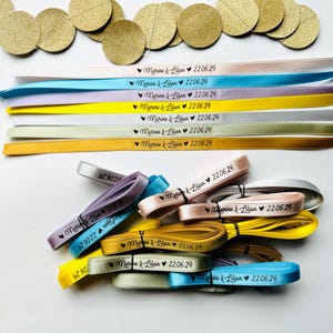 May include: An assortment of colorful ribbons and gold circular confetti. The ribbons are in various colors, including pink, blue, and yellow, and are printed with the text "Marine & Lilian 22.06.29".