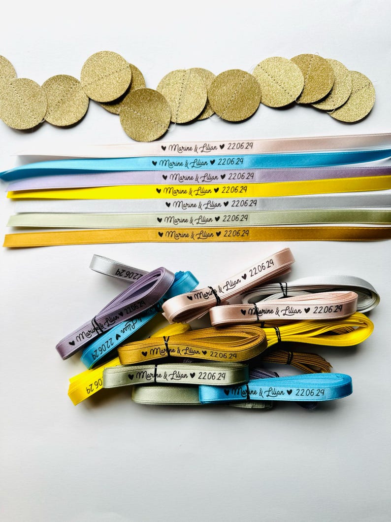 May include: An assortment of colorful ribbons and gold circular confetti. The ribbons are in various colors, including pink, blue, and yellow, and are printed with the text "Marine & Lilian 22.06.29".
