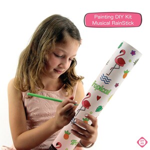May include: A child colors a white cardboard tube with a green marker. The tube has a flamingo and pineapple design and the words "Tropical!" printed on it. The tube is a DIY musical rainstick kit.