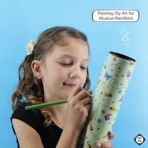 K&ouml;nnte beinhalten: Ein Kind bemalt ein DIY-Set f&uuml;r einen musikalischen Regenstick. Das Set ist eine Pappr&ouml;hre mit einem gr&uuml;nen, gelben und blauen Design aus Schmetterlingen, Bienen und Blumen. Der Text "Painting DIY Kit for Musical RainStick" befindet sich in einer wei&szlig;en Sprechblase oben im Bild.