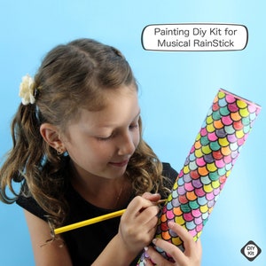 May include: A young person coloring a musical rainstick with a yellow pencil. The rainstick is cylindrical and decorated with a colorful scale pattern. Text on the image reads "Painting DIY Kit for Musical RainStick".