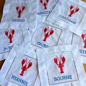 May include: White square linen coasters with a red embroidered lobster design and blue text. The coasters are personalized with various town names, including Dennis, Chatham, and Hyannis. Each coaster has a decorative stitched border.