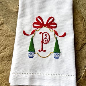 May include: White hand towel with a red bow and the letter "P" embroidered in red. The design includes two green Christmas trees in blue and white pots, surrounded by gold detailing. The towel has a textured surface.