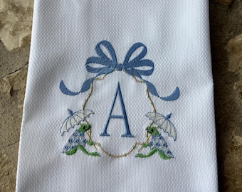 May include: White hand towel with a blue embroidered design. The design includes a blue bow, the letter "A", and two frogs holding umbrellas. The towel has a textured pattern and a decorative stitched border.